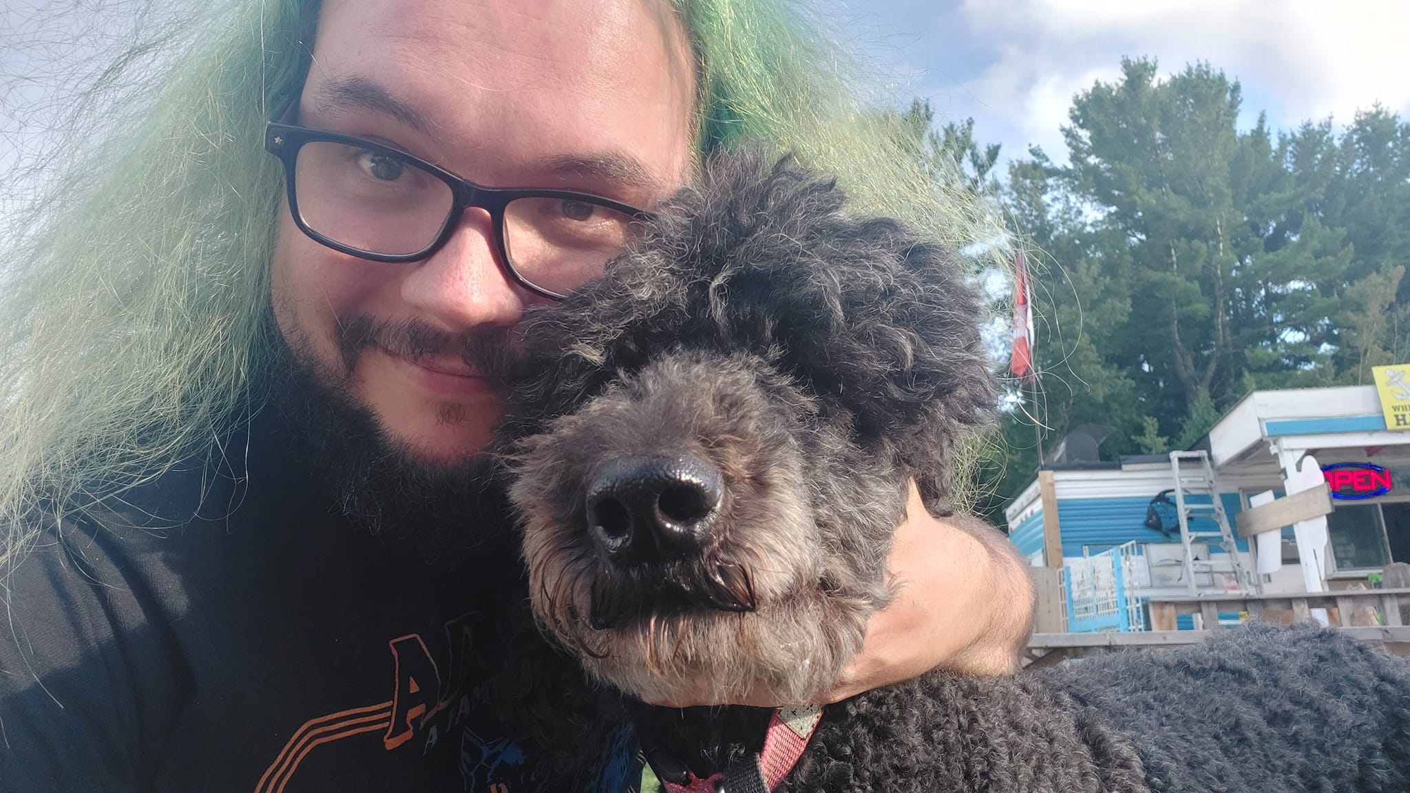 A man with green hair and a beard is hugging a black standard poodle who is too excited for stillness